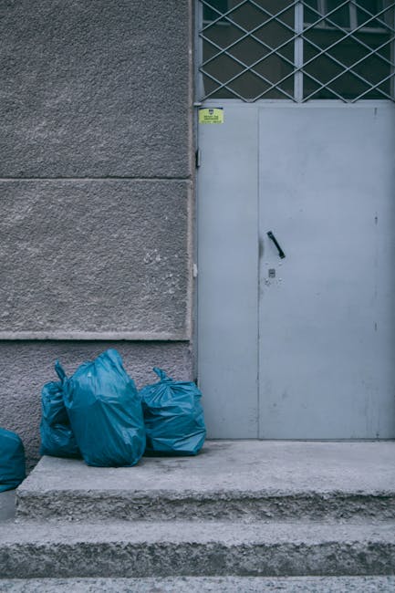 A group of four large blue plastic rubbish bags, tightly tied at the top, are placed on the concrete doorstep of an exterior building. The bags appear filled with waste and are situated next to a smal