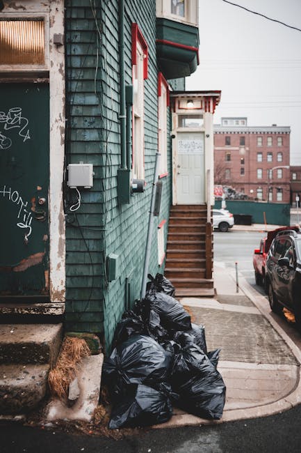 A group of four large blue plastic rubbish bags, tightly tied at the top, are placed on the concrete doorstep of an exterior building. The bags appear filled with waste and are situated next to a smal