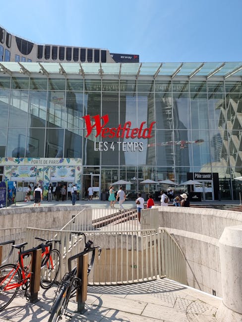A modern glass-fronted shopping centre with the large red 'Westfield' sign and the text 'Les 4 Temps' beneath it. In front of the entrance, there are several pedestrians walking on the paved area, som