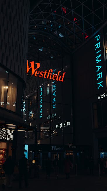 A modern glass-fronted shopping centre with the large red 'Westfield' sign and the text 'Les 4 Temps' beneath it. In front of the entrance, there are several pedestrians walking on the paved area, som